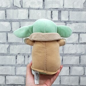 May include: A plush toy of a character with large ears, green skin, and a tan robe. The toy is held in a hand, with a Star Wars tag visible. The background is a white brick wall.