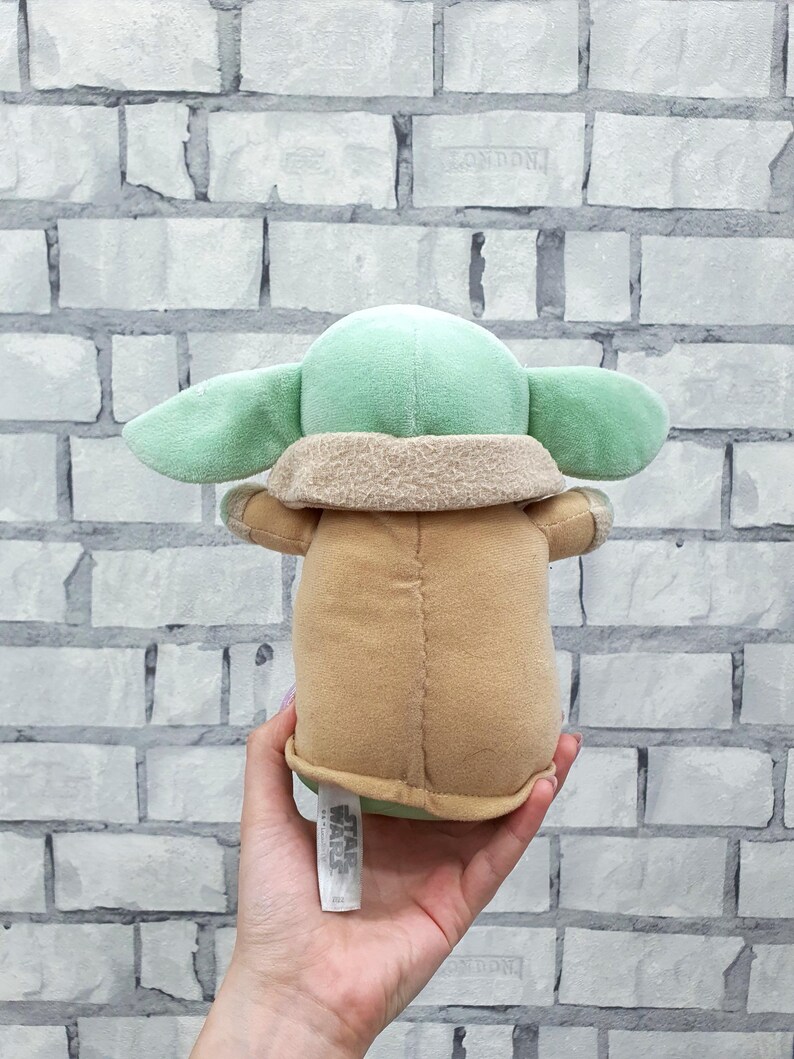 May include: A plush toy of a character with large ears, green skin, and a tan robe. The toy is held in a hand, with a Star Wars tag visible. The background is a white brick wall.