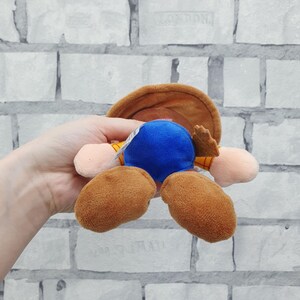 K&ouml;nnte beinhalten: Ein Pl&uuml;schtier, das eine Figur mit braunem Cowboyhut, blauem K&ouml;rper und braunen Stiefeln darstellt. Das Spielzeug wird vor einem wei&szlig;en Backsteinmauer-Hintergrund gehalten.