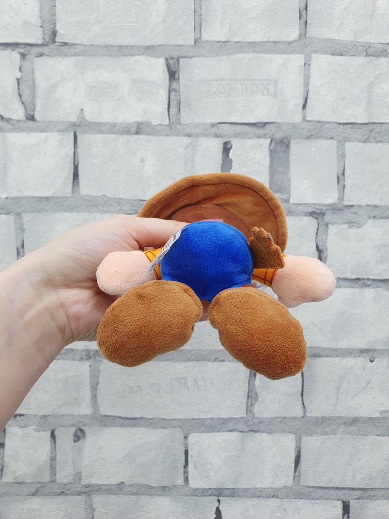 K&ouml;nnte beinhalten: Ein Pl&uuml;schtier, das eine Figur mit braunem Cowboyhut, blauem K&ouml;rper und braunen Stiefeln darstellt. Das Spielzeug wird vor einem wei&szlig;en Backsteinmauer-Hintergrund gehalten.