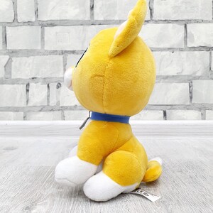 May include: A plush toy dog in a sitting position. The dog is primarily yellow with white paws and a white muzzle. It has a blue collar and a small tail. The toy is set against a light gray brick wall and a light gray wooden surface.