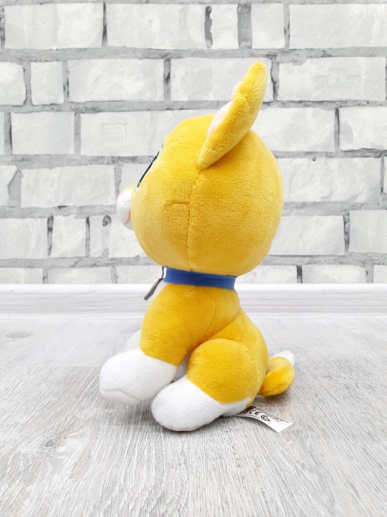 May include: A plush toy dog in a sitting position. The dog is primarily yellow with white paws and a white muzzle. It has a blue collar and a small tail. The toy is set against a light gray brick wall and a light gray wooden surface.