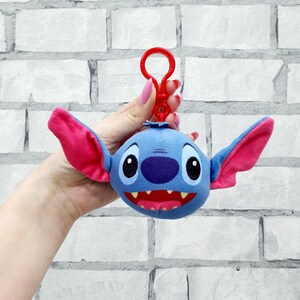 Może przedstawiać: Niebieski pluszowy brelok do kluczy Stitch z r&oacute;żowymi uszami i czerwonymi ustami. Brelok ma czerwony klips. Twarz postaci ma duże czarne oczy i fioletowy nos.