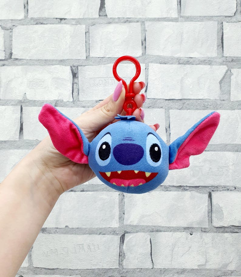 Może przedstawiać: Niebieski pluszowy brelok do kluczy Stitch z r&oacute;żowymi uszami i czerwonymi ustami. Brelok ma czerwony klips. Twarz postaci ma duże czarne oczy i fioletowy nos.