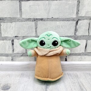 May include: A plush toy of a character with large black eyes, green skin, and large ears. The character is wearing a tan coat with a beige collar. The toy is approximately 8 inches tall.
