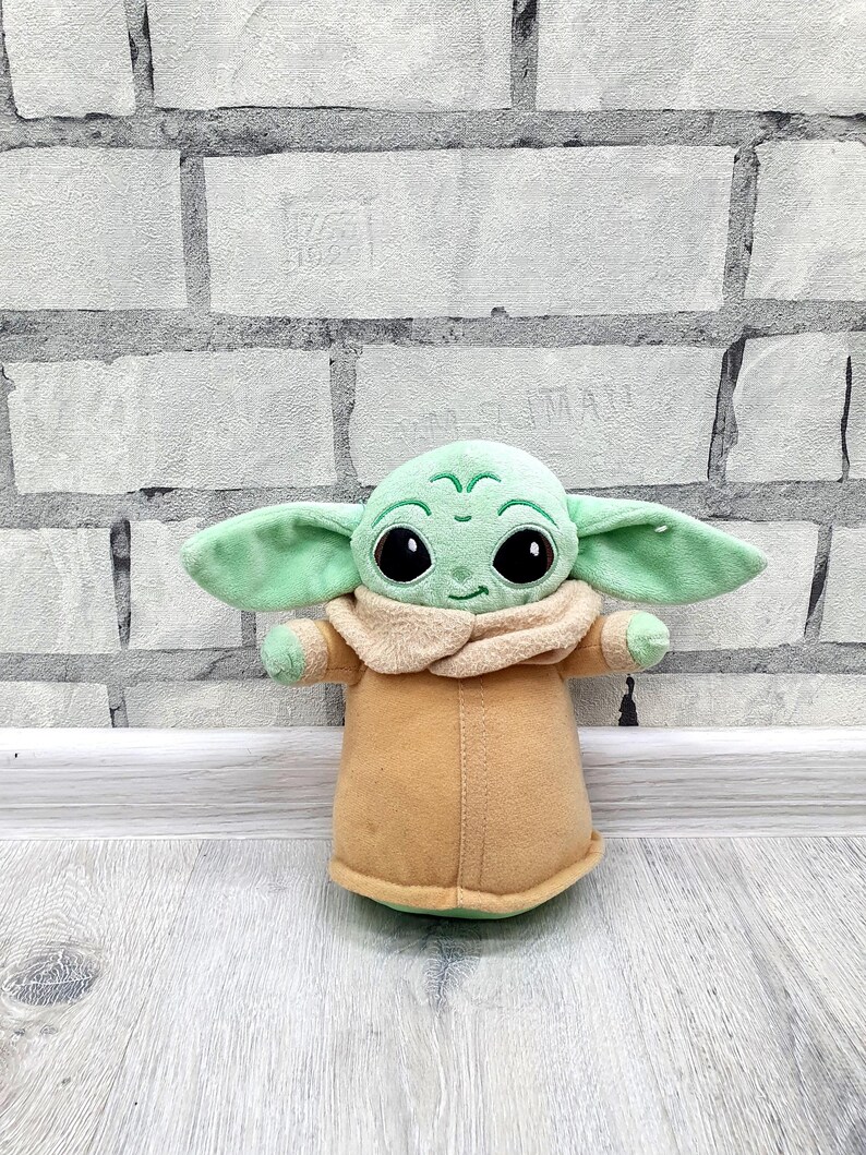 May include: A plush toy of a character with large black eyes, green skin, and large ears. The character is wearing a tan coat with a beige collar. The toy is approximately 8 inches tall.