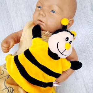 Puede incluir: Una muñeca bebé con un atuendo amarillo y sosteniendo un juguete de peluche de abeja. El juguete de abeja es amarillo con rayas negras y una cara sonriente. La muñeca tiene ojos azules y una expresión neutra.