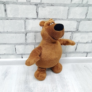 May include: A brown plush bear toy with a black nose and outstretched arms. The stuffed animal has a round body, small ears, and is standing upright. The bear is made of soft, fuzzy material.