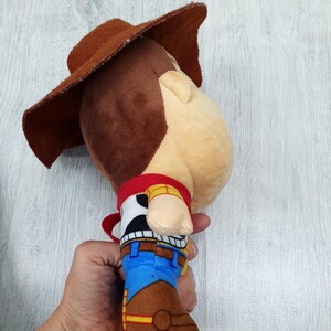 May include: A soft toy cowboy character, with a brown hat, red neckerchief, and a blue and brown outfit. The toy has a light skin tone and a stitched design, resembling a well-known animated character.