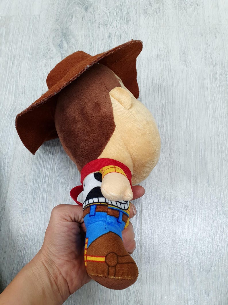 May include: A soft toy cowboy character, with a brown hat, red neckerchief, and a blue and brown outfit. The toy has a light skin tone and a stitched design, resembling a well-known animated character.
