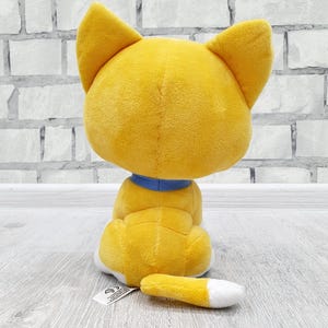 May include: A plush toy cat, viewed from the back, in a sitting position. The cat is primarily yellow with a blue collar and a white-tipped tail. The toy is on a light gray surface with a brick wall background.