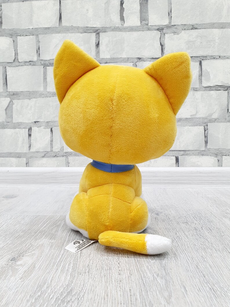 May include: A plush toy cat, viewed from the back, in a sitting position. The cat is primarily yellow with a blue collar and a white-tipped tail. The toy is on a light gray surface with a brick wall background.