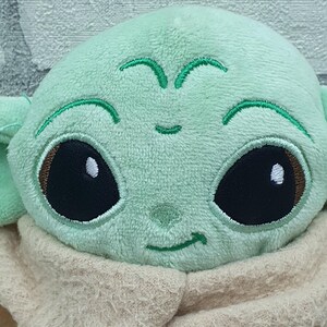 May include: A plush toy featuring a character with large black eyes, a light green face, and large ears. The character is wearing a tan-colored robe. The toy is made of soft, plush material.