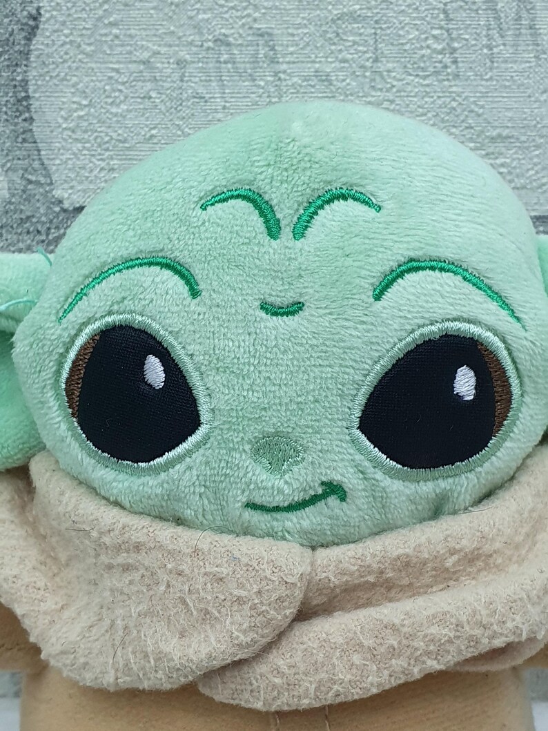 May include: A plush toy featuring a character with large black eyes, a light green face, and large ears. The character is wearing a tan-colored robe. The toy is made of soft, plush material.