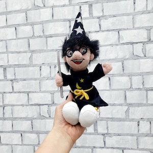 Puede incluir: Muñeco de mago de peluche con una túnica negra, un sombrero puntiagudo con estrellas y gafas redondas. El muñeco tiene una cara sonriente, una luna creciente y una estrella amarillas en la túnica y sostiene una varita plateada.