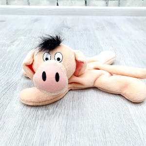 May include: A plush pig toy in a light peach color, lying on a light gray surface. The pig has a pink snout with two black nostrils, large cartoon eyes, and a tuft of black hair. The toy's ears are also pink.