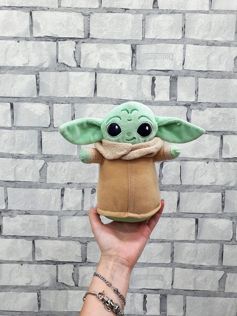 May include: A plush toy of a character with large eyes, green skin, and large ears. The character is wearing a tan coat and a beige collar. The toy is being held in a person's hand against a white brick wall.