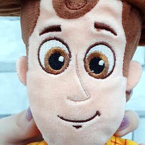 K&ouml;nnte beinhalten: Ein Pl&uuml;schtier einer Figur mit braunem Cowboyhut, braunen Haaren und einem gelben karierten Hemd. Die Figur hat gro&szlig;e, gestickte braune Augen und ein aufgen&auml;htes L&auml;cheln. Das Spielzeug wird von einer Hand mit lackiertem Fingernagel gehalten.