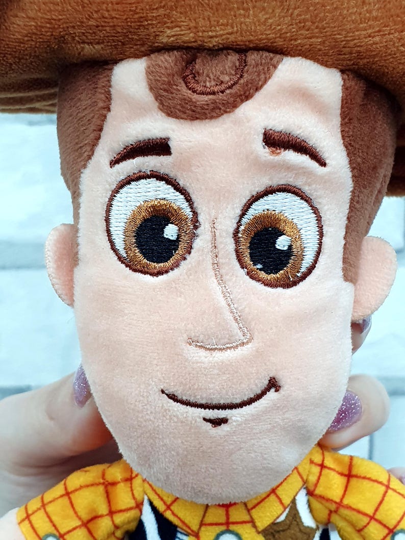 K&ouml;nnte beinhalten: Ein Pl&uuml;schtier einer Figur mit braunem Cowboyhut, braunen Haaren und einem gelben karierten Hemd. Die Figur hat gro&szlig;e, gestickte braune Augen und ein aufgen&auml;htes L&auml;cheln. Das Spielzeug wird von einer Hand mit lackiertem Fingernagel gehalten.