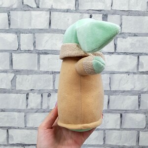 May include: A plush toy of a character with a large head and pointed ears. The toy has a light green head, tan robe, and small arms. The toy is held in a hand against a white brick wall.
