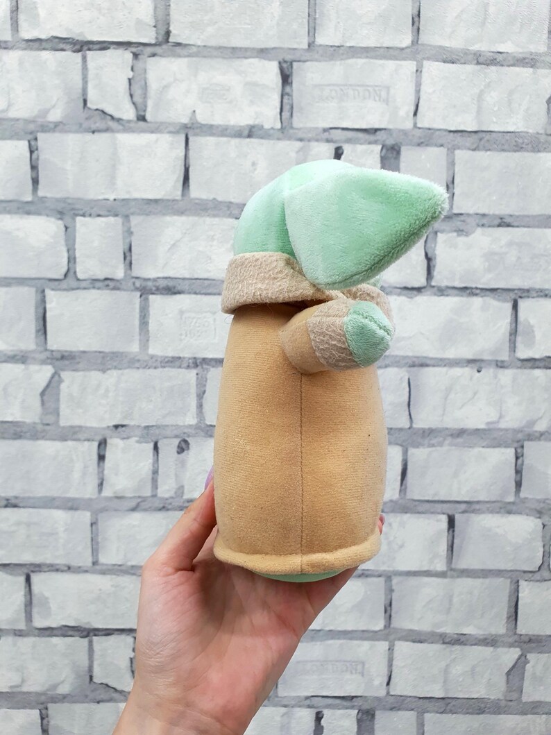 May include: A plush toy of a character with a large head and pointed ears. The toy has a light green head, tan robe, and small arms. The toy is held in a hand against a white brick wall.