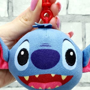 Może przedstawiać: Niebieski pluszowy brelok do kluczy Stitch z dużymi czarnymi oczami, fioletowym nosem i szerokimi czerwonymi ustami z ż&oacute;łtymi zębami. Brelok ma r&oacute;żowe wewnętrzne uszy i czerwony plastikowy klips.