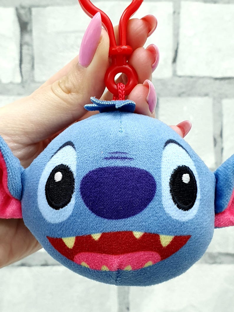Może przedstawiać: Niebieski pluszowy brelok do kluczy Stitch z dużymi czarnymi oczami, fioletowym nosem i szerokimi czerwonymi ustami z ż&oacute;łtymi zębami. Brelok ma r&oacute;żowe wewnętrzne uszy i czerwony plastikowy klips.