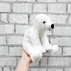 May include: A white plush polar bear toy with a black nose and small black eyes. The stuffed animal is sitting and has a soft, fluffy texture. The background is a white brick wall.