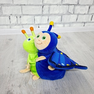 Può includere: Due peluche: una farfalla blu e un bruco verde. La farfalla ha grandi ali blu con dettagli blu scuro e gialli. Il bruco è verde lime con antenne arancioni. Entrambi i giocattoli hanno facce sorridenti.