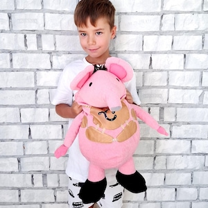 Puede incluir: Un juguete de elefante de peluche rosa con orejas grandes, pies negros y un diseño de parche marrón. El juguete lo sostiene una persona que lleva una camisa blanca y pantalones cortos con un estampado de coche. El fondo es una pared de ladrillos blancos.