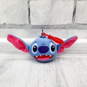 Może przedstawiać: Pluszowy brelok do kluczy z postacią Stitcha. Niebieskie stworzenie o okrągłej twarzy ma duże r&oacute;żowe uszy, fioletowy nos i szeroki, zębaty uśmiech. Do g&oacute;ry przymocowany jest czerwony plastikowy klips. Zabawka jest ustawiona na tle białej ceglanej ściany i jasnej drewnianej powierzchni.