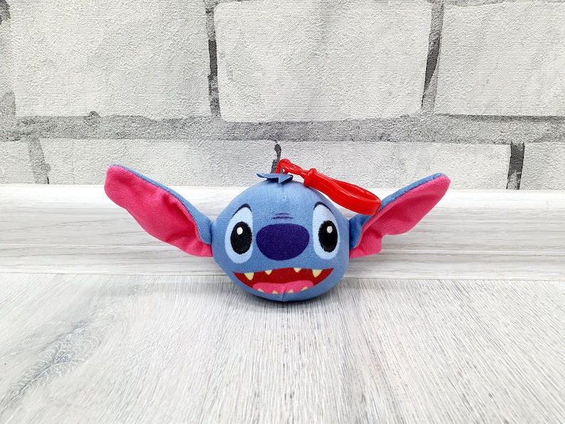 Może przedstawiać: Pluszowy brelok do kluczy z postacią Stitcha. Niebieskie stworzenie o okrągłej twarzy ma duże r&oacute;żowe uszy, fioletowy nos i szeroki, zębaty uśmiech. Do g&oacute;ry przymocowany jest czerwony plastikowy klips. Zabawka jest ustawiona na tle białej ceglanej ściany i jasnej drewnianej powierzchni.