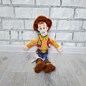 May include: A soft toy cowboy character, wearing a brown hat, a yellow plaid shirt, a red waistcoat with a Christmas tree design, blue jeans, and brown boots. The toy has a cheerful expression and outstretched arms.