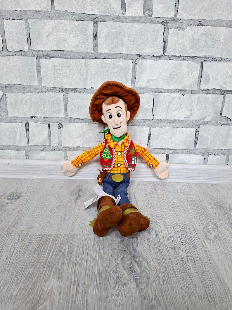 May include: A soft toy cowboy character, wearing a brown hat, a yellow plaid shirt, a red waistcoat with a Christmas tree design, blue jeans, and brown boots. The toy has a cheerful expression and outstretched arms.