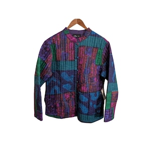 May include: A quilted jacket with a mandarin collar and button-up front. The jacket features a patchwork design in shades of purple, teal, green, and magenta. The jacket is hanging on a wooden hanger.