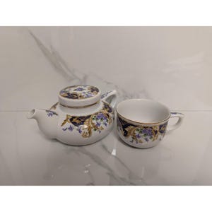 May include: White porcelain teapot with lid and matching teacup. Both pieces feature a decorative design with navy blue, gold, and purple floral patterns. The teapot has a curved spout and handle, while the teacup has a small handle.