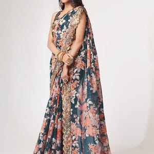 May include: A dark teal saree with a floral pattern in shades of peach and white. The saree features a gold-colored border with intricate detailing. The fabric drapes elegantly, showcasing the floral design.