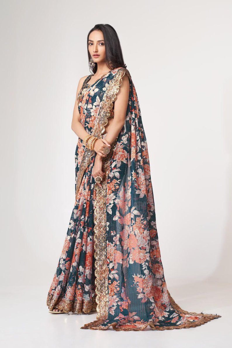 May include: A dark teal saree with a floral pattern in shades of peach and white. The saree features a gold-colored border with intricate detailing. The fabric drapes elegantly, showcasing the floral design.