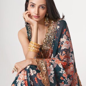 May include: A woman wearing a dark teal sari with a floral pattern in shades of orange and white. The sari is trimmed with gold lace and sequins. She is wearing gold bangles, earrings, and rings.