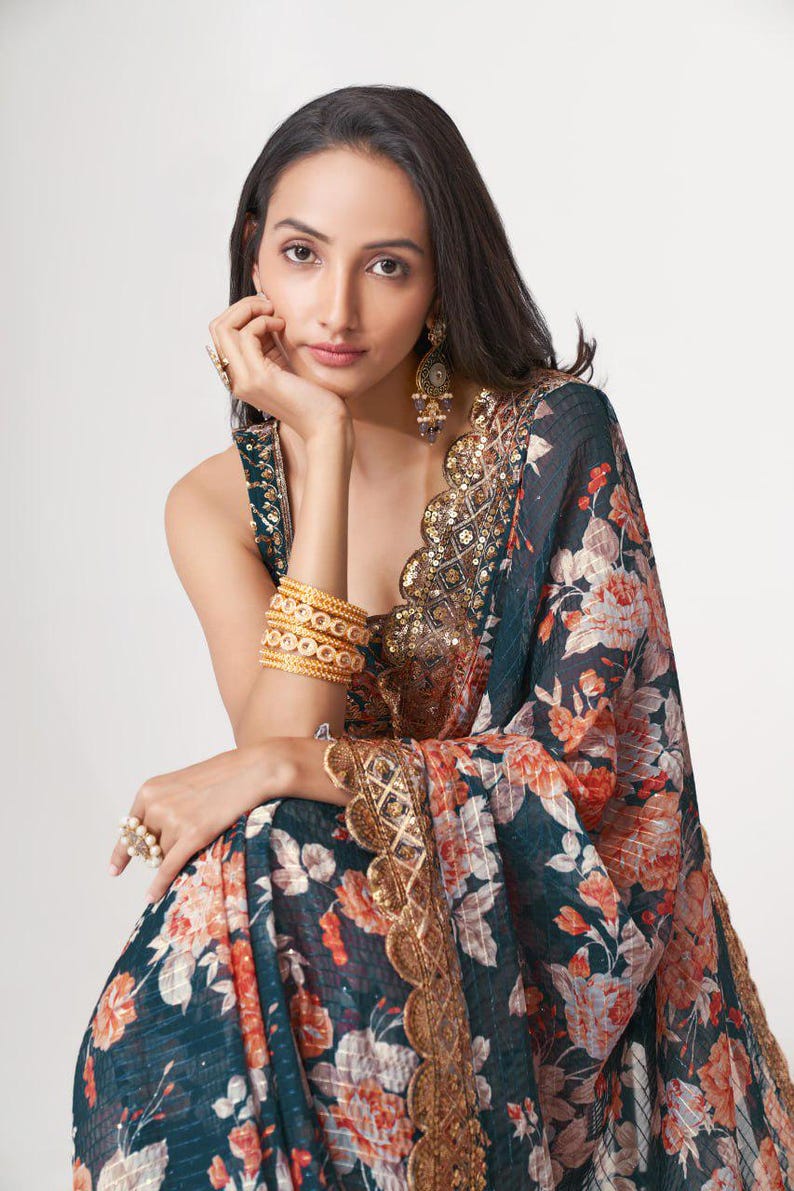 May include: A woman wearing a dark teal sari with a floral pattern in shades of orange and white. The sari is trimmed with gold lace and sequins. She is wearing gold bangles, earrings, and rings.