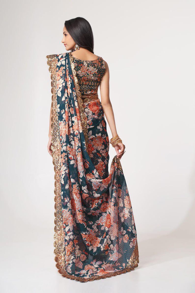 May include: A dark teal saree with a floral pattern in shades of orange and pink. The saree features a gold sequined border and is paired with a matching blouse. The overall design is elegant and suitable for special occasions.