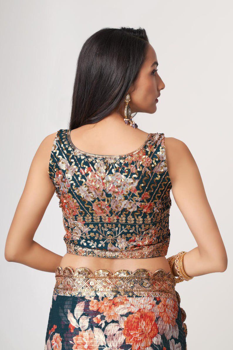 May include: A sleeveless, dark teal top with intricate floral embroidery in shades of orange, gold, and pink. The top features a detailed sequin design and is paired with a matching floral skirt. A gold bracelet and earrings complete the look.