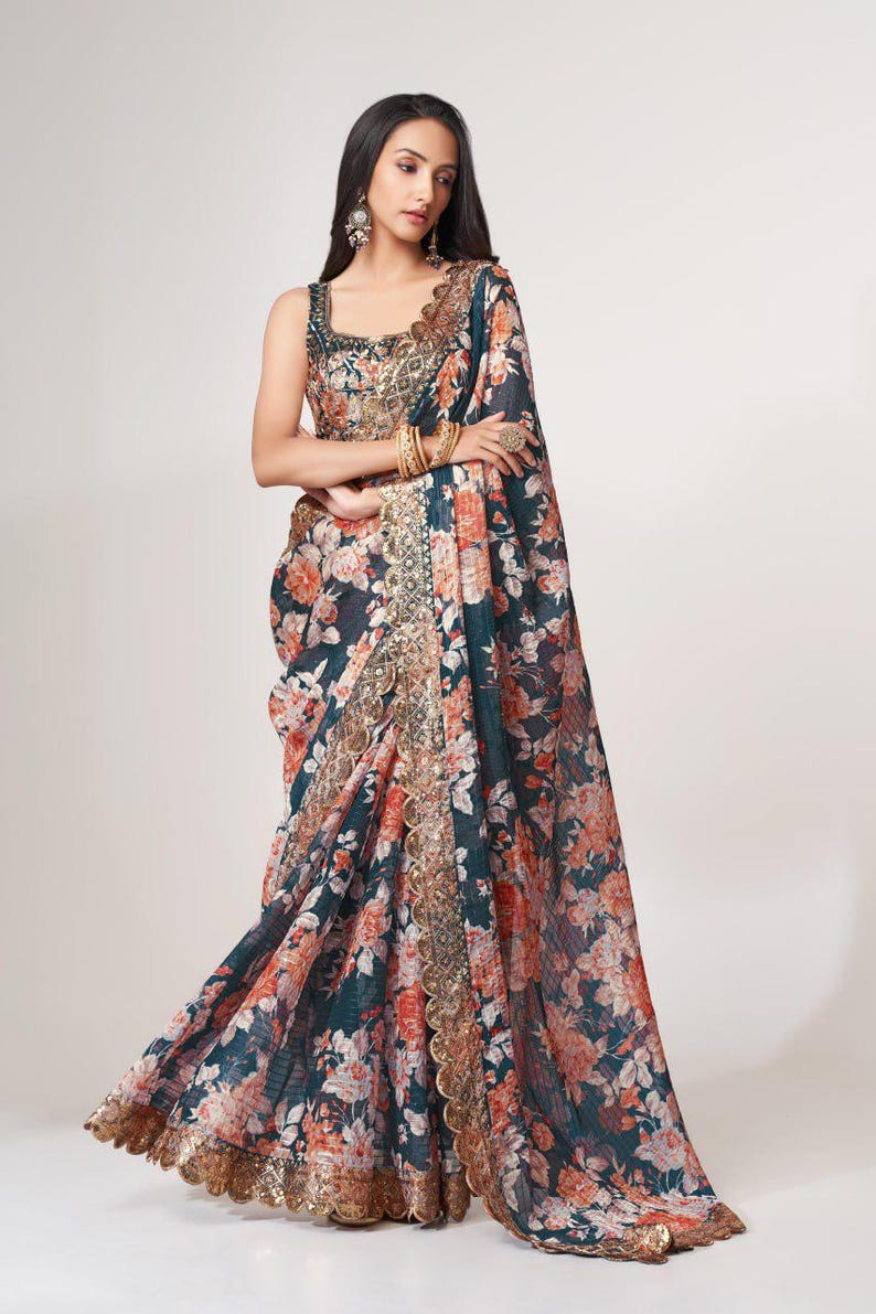 May include: A flowing, floral-print saree in shades of teal, coral, and cream. The saree features a detailed gold border and matching sleeveless blouse. The fabric drapes elegantly, showcasing the intricate floral pattern. A traditional Indian garment.