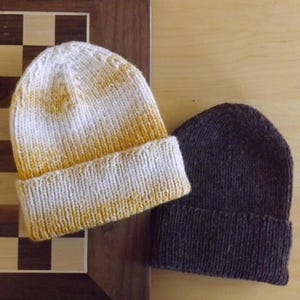May include: Two knitted beanies. One is white with a yellow gradient, and the other is a solid dark grey. Both have a folded brim. The beanies are displayed on a wooden surface, with a portion of a chessboard visible.