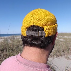 May include: A mustard yellow knit beanie cap with a rolled brim and a grey fabric strap featuring a metallic buckle at the back. The cap has a textured, hand-knitted appearance and is designed to sit snugly on the head with an adjustable closure.