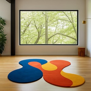 Puede incluir: Una alfombra abstracta con una forma inusual, en colores azul, naranja y amarillo. Se encuentra sobre un suelo de madera en una habitación con una gran ventana que da a un paisaje verde y arbolado.