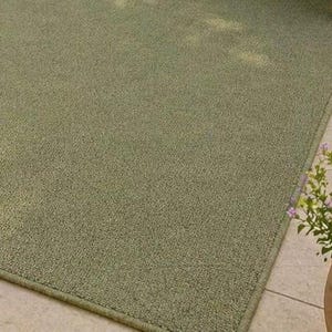 May include: A rectangular, sage green outdoor rug with a textured surface. The rug has a bound edge and is placed on a tiled surface. A potted plant with purple flowers is in the corner.
