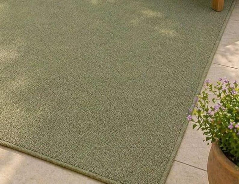 May include: A rectangular, sage green outdoor rug with a textured surface. The rug has a bound edge and is placed on a tiled surface. A potted plant with purple flowers is in the corner.