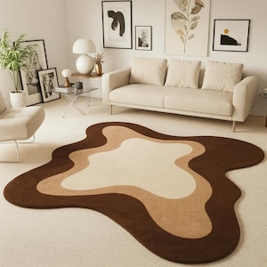 Puede incluir: Una alfombra de &aacute;rea marr&oacute;n, beige y crema con una forma org&aacute;nica y fluida. La alfombra est&aacute; colocada sobre una alfombra de color claro en un sal&oacute;n, con un sof&aacute; beige y obras de arte en la pared.