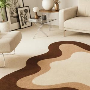 Puede incluir: Un sal&oacute;n moderno con una alfombra color crema con un dise&ntilde;o ondulado en tonos marr&oacute;n y beige. La habitaci&oacute;n incluye un sof&aacute; beige, una mesa auxiliar de cristal y obras de arte enmarcadas. Una planta en maceta y objetos decorativos completan la est&eacute;tica de la habitaci&oacute;n.